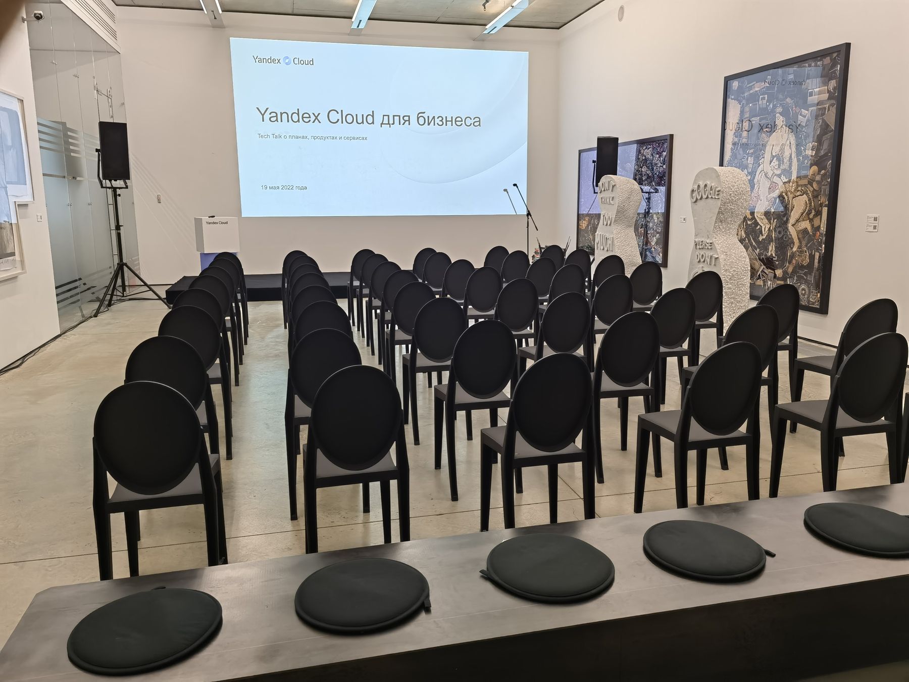 Зал галереи перед началом: экран «Yandex Cloud для бизнеса. Tech Talk о планах, продуктах и сервисах», кафедра, ряды стульев среди работ экспозиции, 19.05.2022