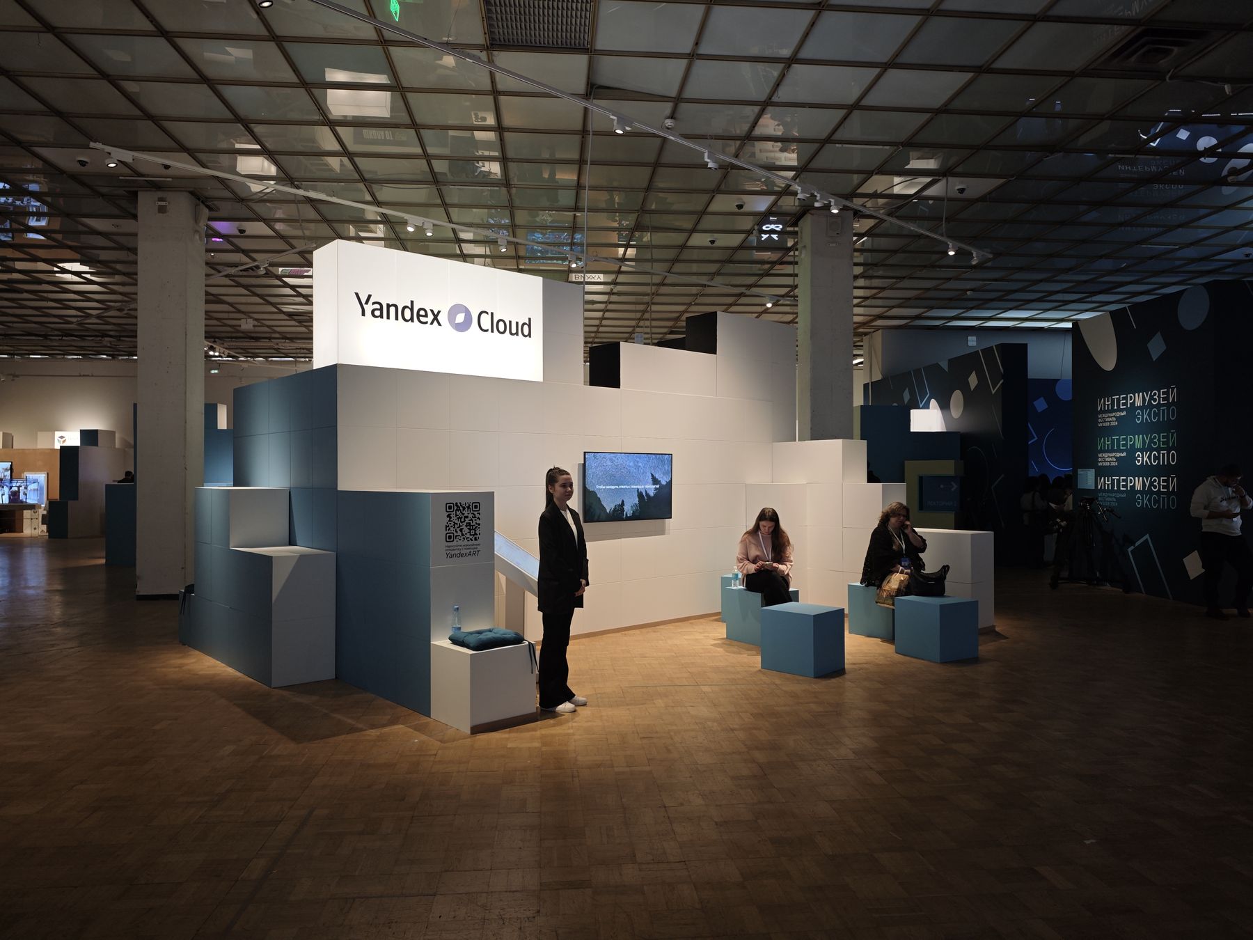 Стенд Yandex Cloud на «Интермузей Экспо»: модульная конструкция из белых и бирюзовых кубов, экран с роликом Центра технологий для общества, QR-код «нарисуйте новогоднюю открытку с помощью YandexART», зона отдыха, 19.12.2024